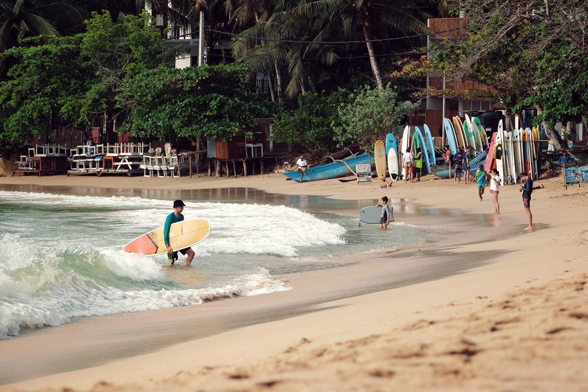 Un surfista que sale del mar con su tabla de surf amarillo y naranja en la arena dorada en la playa de Hiriketiya, Sri Lanka.